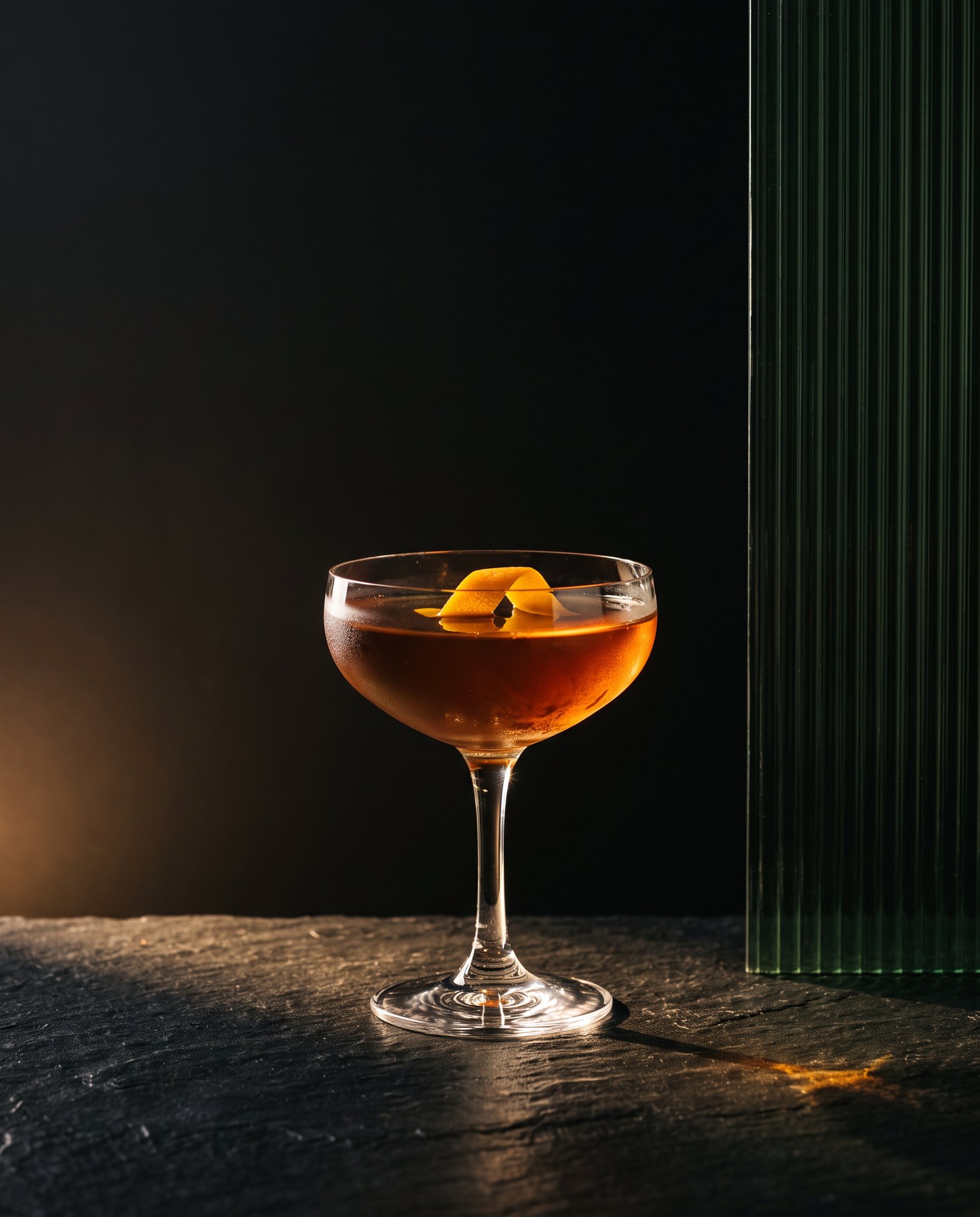 A coupe glass of amber cocktail with a citrus twist on black stone, lit by warm gold light