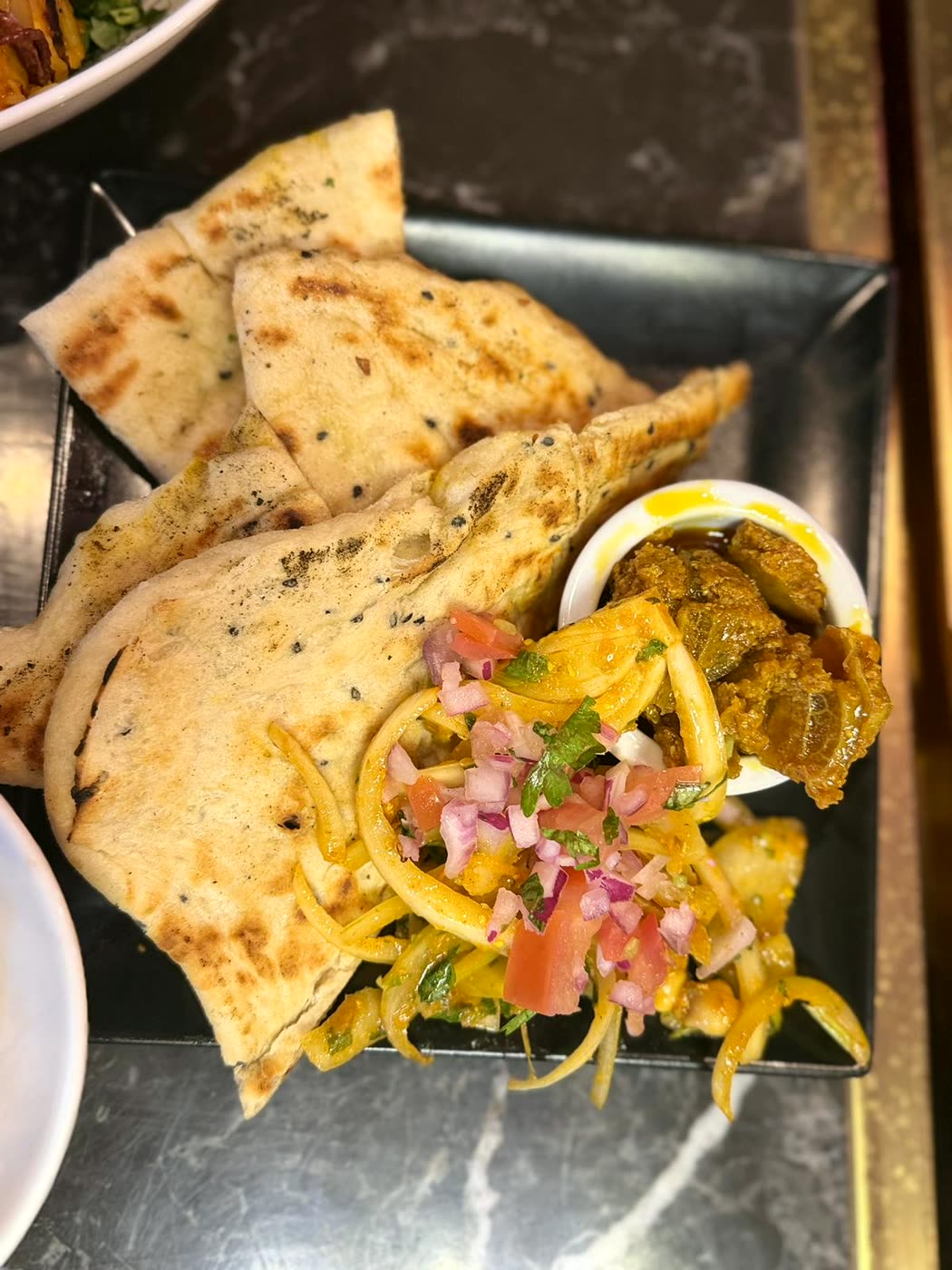 Charred flatbread torn into pieces on a black plate, with a small pot of pickle and a fresh onion, tomato and coriander salad