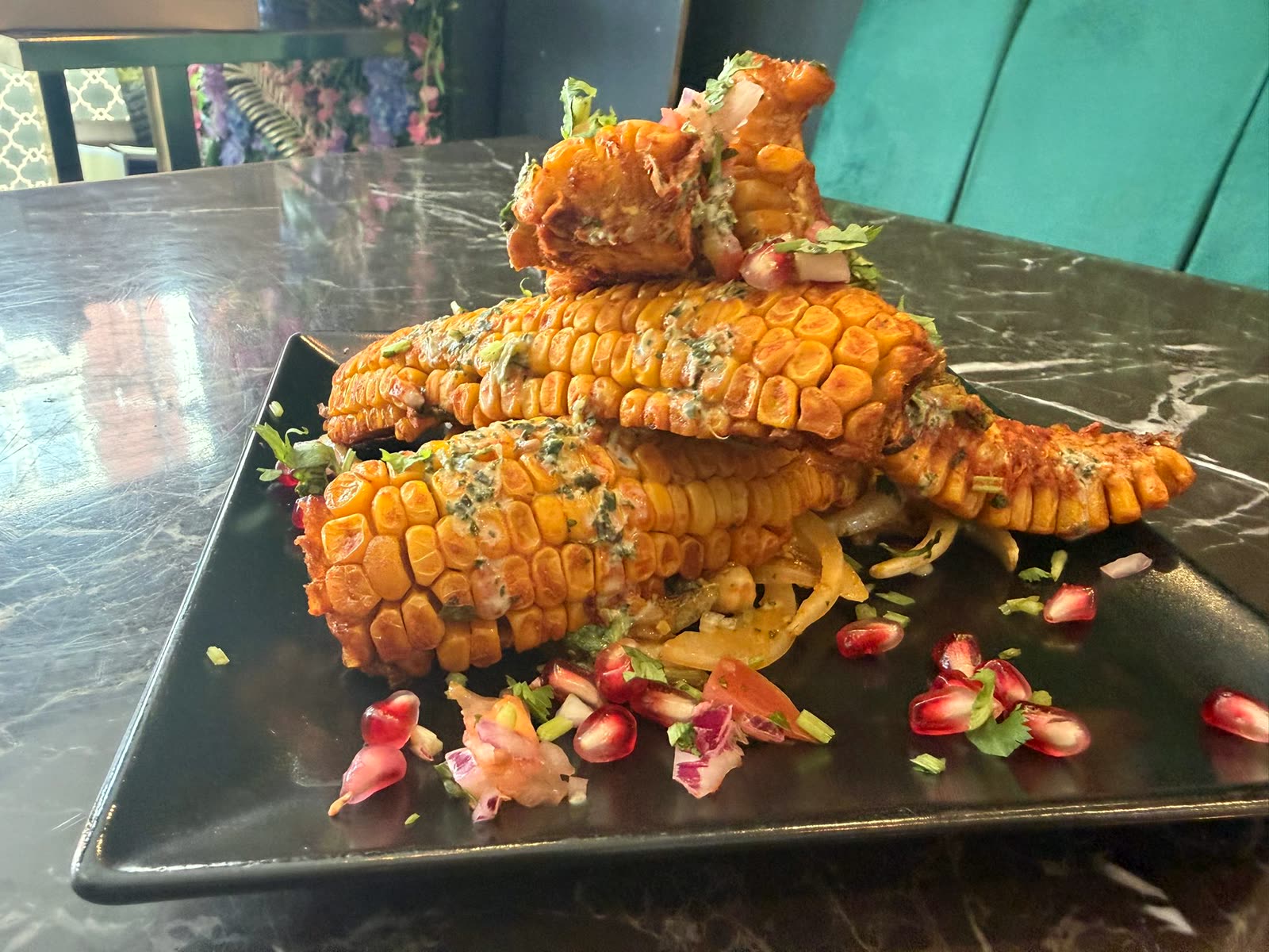 Charred corn ribs topped with herbs, pomegranate seeds and chopped salad on a black plate, on a marble table in HAVN's dining room with green velvet seating behind