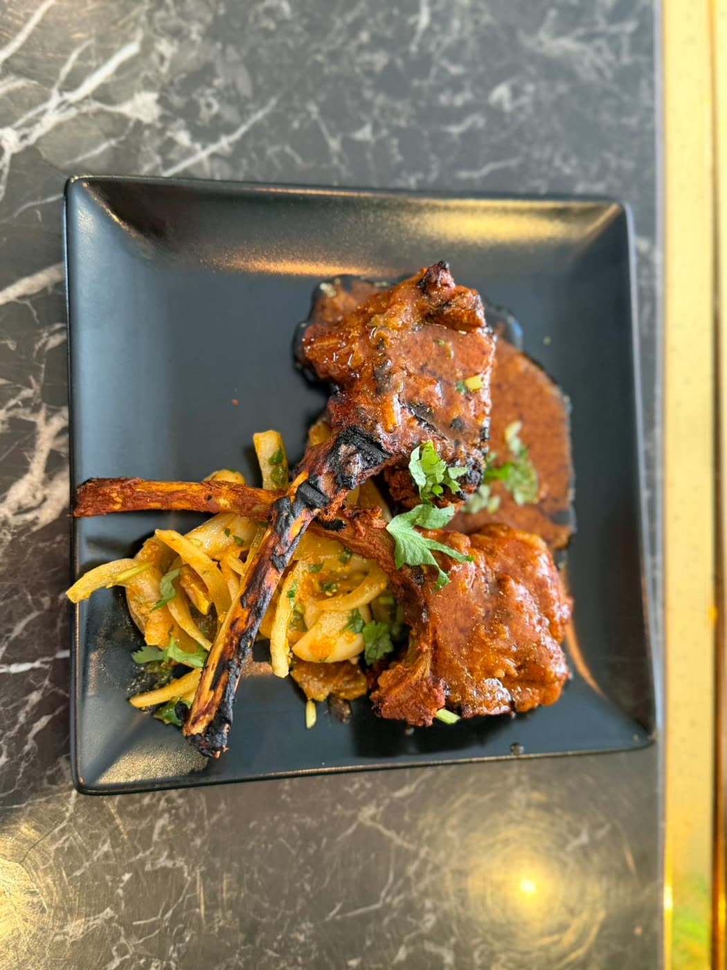 Grilled chops on the bone in a dark spiced sauce, served on a black plate with fried onions and coriander