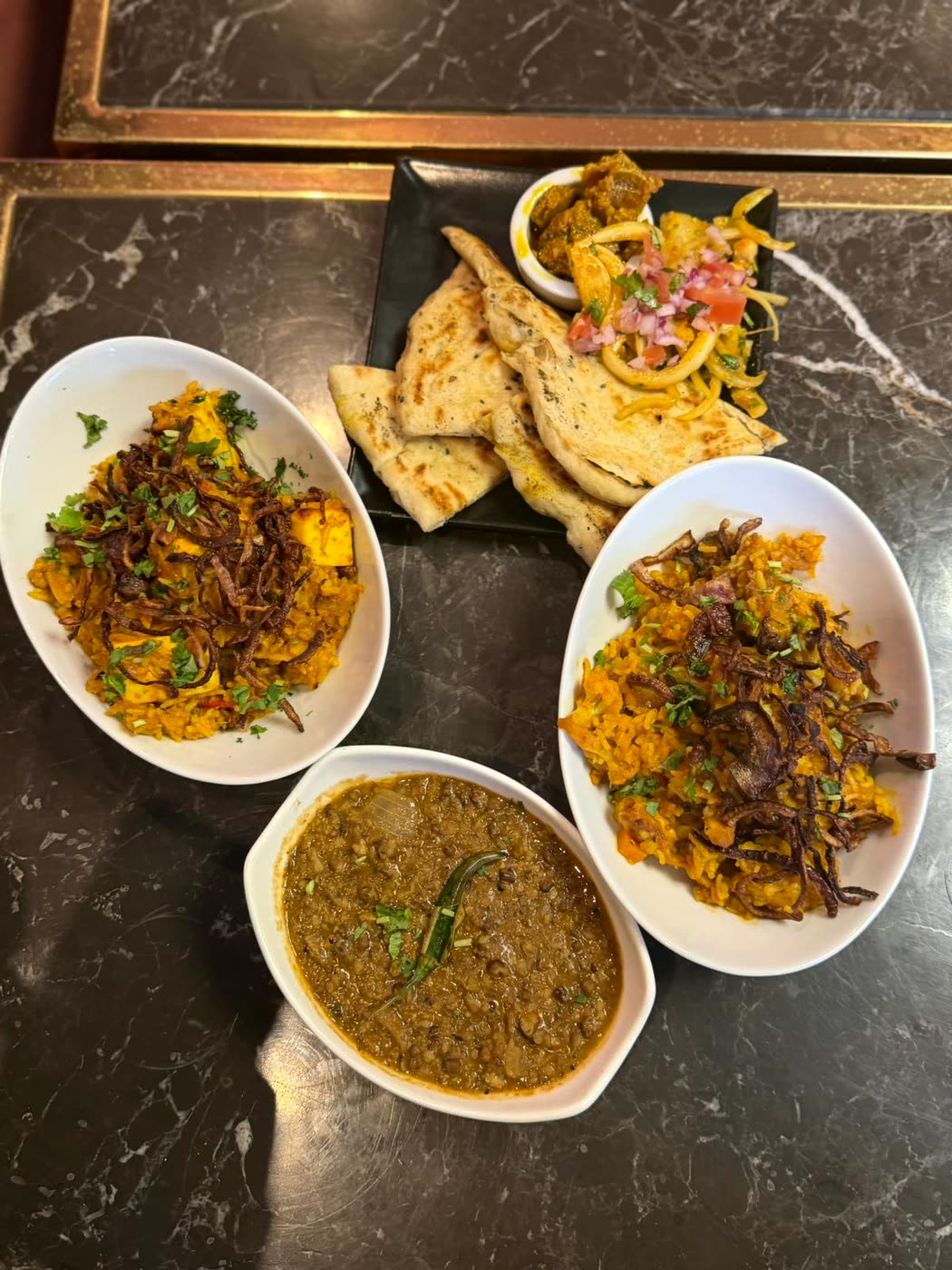 Looking down on a marble table set with two dishes of rice topped with crisp fried onion, a bowl of dark lentil curry and a black plate of flatbread and salad